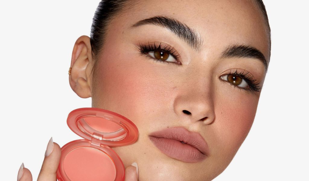 Eine Frau mit dunklen Features hält einen pfirsichfarbenen Blush vor weißem Hintergrund.