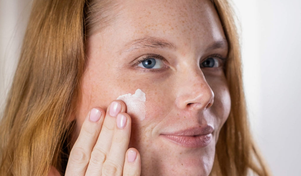 Eine Frau mit rotem Haar und blauen Augen lächelt, während sie Creme auf ihre Wange trägt.