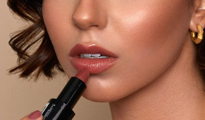 Nahaufnahme einer Frau, die glänzenden Lippenstift in Rosé aufträgt, mit dezentem Make-up und Goldohrring.