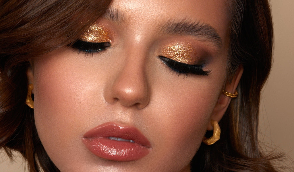Frau mit goldenem Glitzerlidschatten, langen Wimpern und glänzendem Lippenstift in Nahaufnahme.