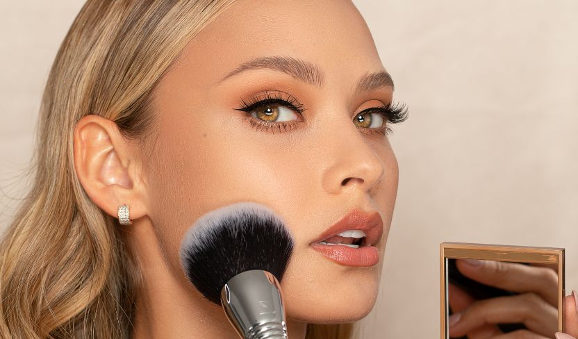 Frau trägt mit einem großen Make-up-Pinsel Puder auf ihre Wange auf und hält einen Kompaktspiegel in der Hand.