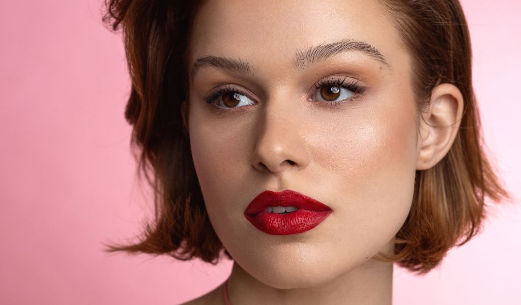Frau mit kurzem braunem Haar, makellosem Teint und auffälligem roten Lippenstift vor rosa Hintergrund, elegant geschminkt.