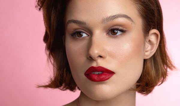 Frau mit kurzem braunem Haar, makellosem Teint und auffälligem roten Lippenstift vor rosa Hintergrund, elegant geschminkt.
