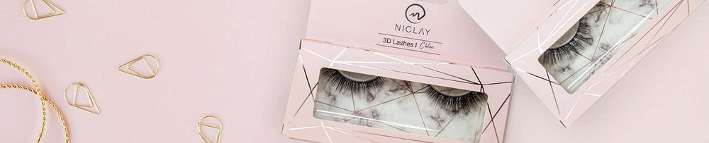Wimpern von NICLAY in rosa Verpackung auf pastellfarbenem Hintergrund mit goldenen Accessoires.