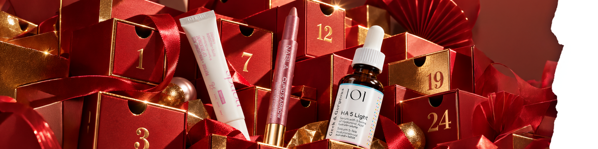 Ein Lippenpflegeprodukt, ein Lippenstift und eine Flasche Gesichtsserum, angeordnet vor gestapelten roten Weihnachtsgeschenkboxen.