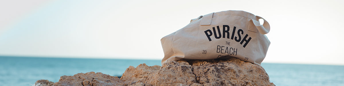 Beige Einkaufstasche mit dem Text „PURISH THE BEACH“ auf Felsen am Meer.