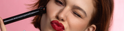 Frau mit rotem Lippenstift zwinkert, während sie vor einem rosa Hintergrund ein schwarzes Make-up-Tool an ihre Wange hält.