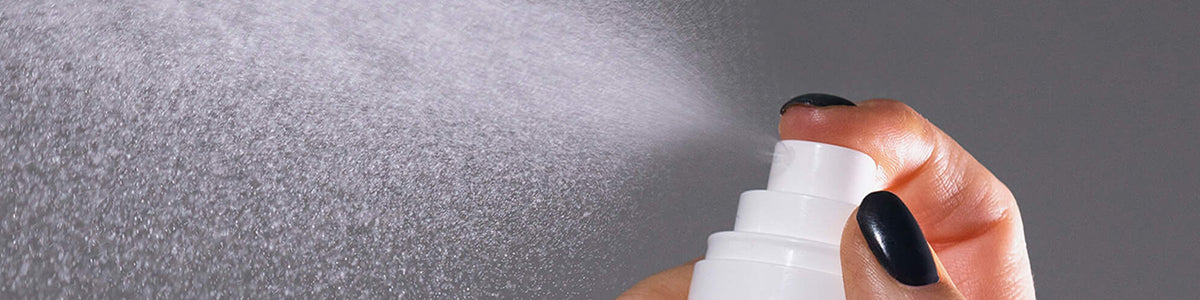 Eine Hand mit dunklen Nägeln sprüht einen feinen Nebel aus einer weißen Sprühflasche vor einem grauen Hintergrund.