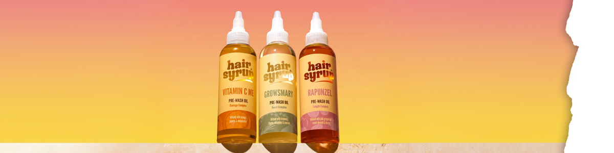 Drei Flaschen Hair Syrup-Öl zur Anwendung vor dem Waschen in den Varianten Vitamin C Me, Growsmary und Rapunzel, angeordnet auf einer Leiste vor einem Hintergrund mit einem Farbverlauf in Sonnenuntergangsfarben.