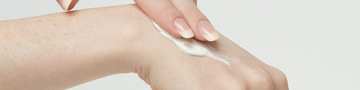 Eine Hand trägt vor einem schlichten weißen Hintergrund weiße Lotion auf den Handrücken einer anderen Hand auf.