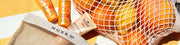 Nuxe-Sonnenschutztuben, ein Netzbeutel mit Orangen und ein Nuxe-Beutel auf gelb-weiß gestreiftem Untergrund.