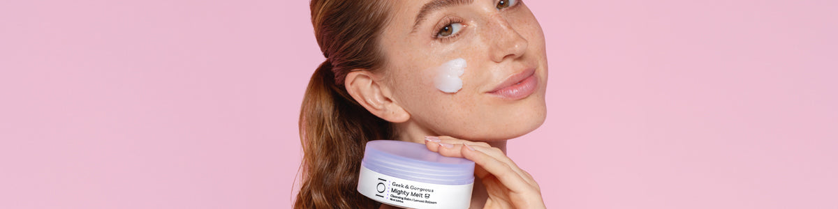 Frau mit frischer Haut trägt Creme auf ihre Wange auf und hält eine Feuchtigkeitscreme von Geek&Gorgeous in der Hand vor einem rosa Hintergrund.