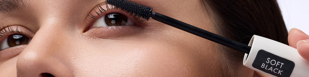 Nahaufnahme eines Auges, auf das gerade schwarze Mascara mit der Aufschrift „Soft Black“ aufgetragen wird.