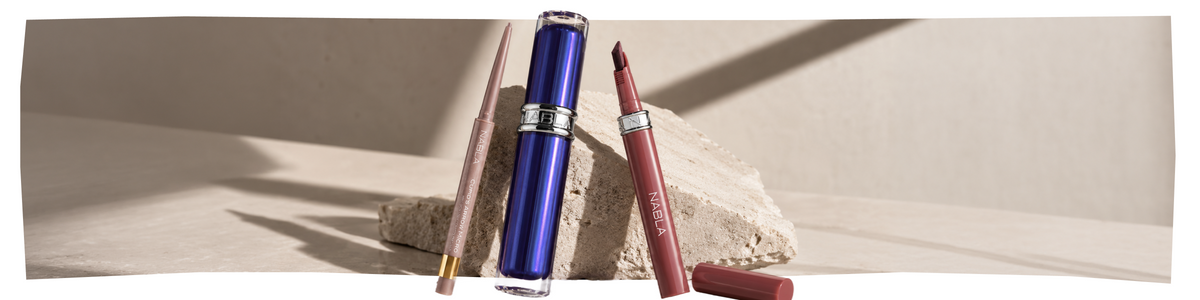 Drei Make-up-Produkte – eine blaue Tube, ein rosa Stift und ein mauvefarbener Lippenstift – sind auf strukturiertem Stein vor einem neutralen beigen Hintergrund mit sanften Schatten angeordnet.
