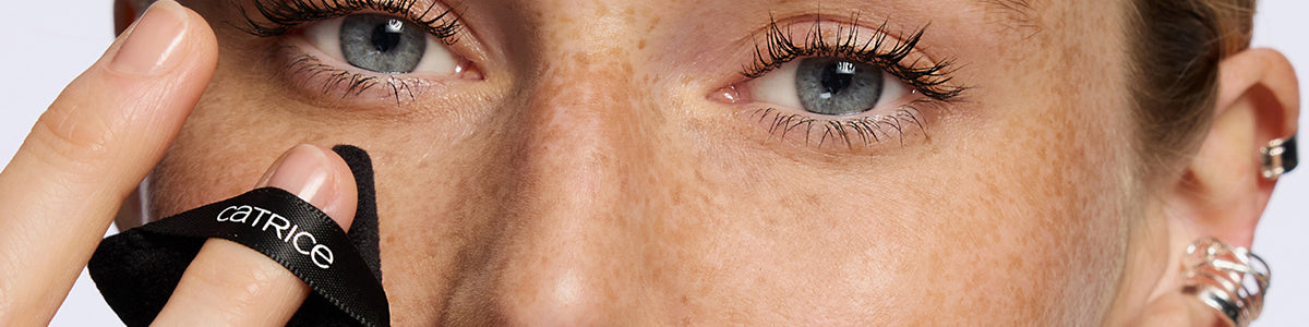 Nahaufnahme eines Gesichts mit blauen Augen und Sommersprossen; eine Frau trägt mit einem schwarzen Catrice-Make-up-Puff Puder auf.