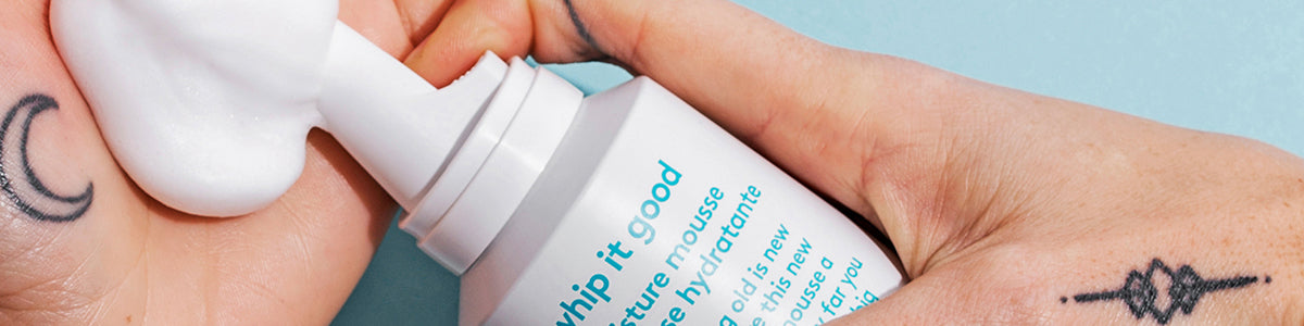 Hand die vor blauem Hintergrund weißen Schaum aus einer Flasche mit der Aufschrift „Whip It Good Moisture Mousse“ ausgibt.