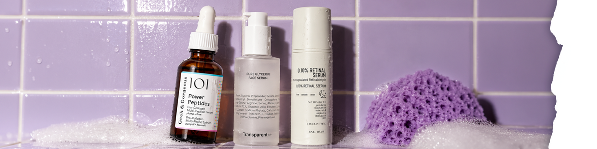 Drei Hautpflegeflaschen – ein Serum, ein Glycerin-Gesichtsserum und ein Retinal-Serum – stehen auf einer nassen, violett gefliesten Fläche neben einem violetten Schwamm.