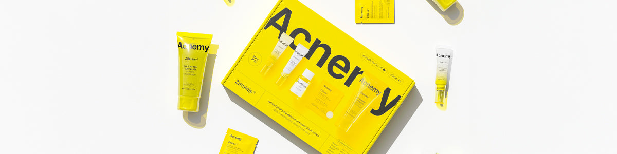 Gelbe Verpackung des Acne-Set „Acnemy“ mit verschiedenen Pflegeprodukten gegen Unreinheiten auf weißem Hintergrund, Produkte locker um die Box arrangiert.