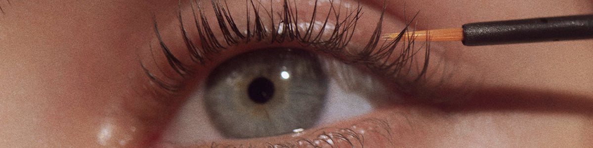 Nahaufnahme eines blauen Auges mit einem Pinsel, der ein klares Produkt auf die oberen Wimpern aufträgt.