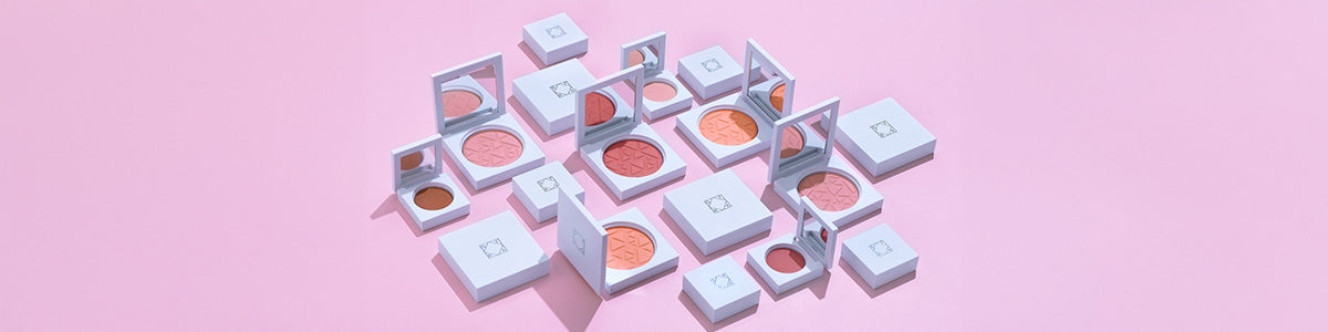 Make-up-Produkte in weißer Verpackung mit Spiegel, darunter Rouge und Bronzer in verschiedenen Nuancen, arrangiert auf rosa Hintergrund.