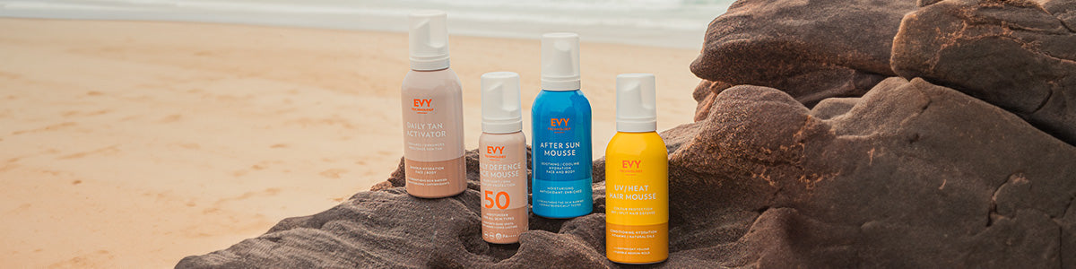 Vier Sonnenschutz- und Bräunungsprodukte auf einem Felsen am Strand, mit Sand und Meer im Hintergrund.