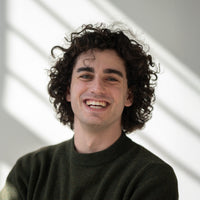 Porträt von André mit dunkelbraunen Locken, braunen Augen und dunkelgrünem Oberteil vor hellem Hintergrund.