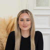  Porträt von Sara mit blonden Haaren und schwarzem Oberteil vor hellem Hintergrund.
