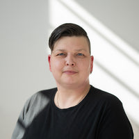 Porträt von Nancy mit dunkelbraunen Haaren, blauen Augen und schwarzem Oberteil vor hellem Hintergrund.