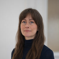 Porträt von Sylvia mit braunen Haaren, blauen Augen und schwarzem Oberteil vor hellem Hintergrund.
