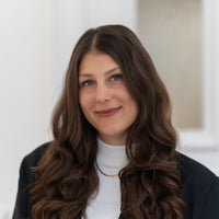 Porträt von Carina mit braunen Haaren, blauen Augen und schwarzem Blazer vor hellem Hintergrund.