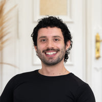 Porträt von Gianluca mit schwarzen Haaren, braunen Augen und schwarzem Oberteil vor hellem Hintergrund.