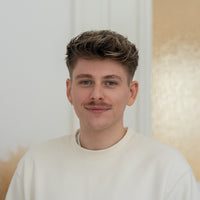 Porträt von Ron mit dunkelblonden Haaren, grünen Augen und beigem Oberteil vor hellem Hintergrund.