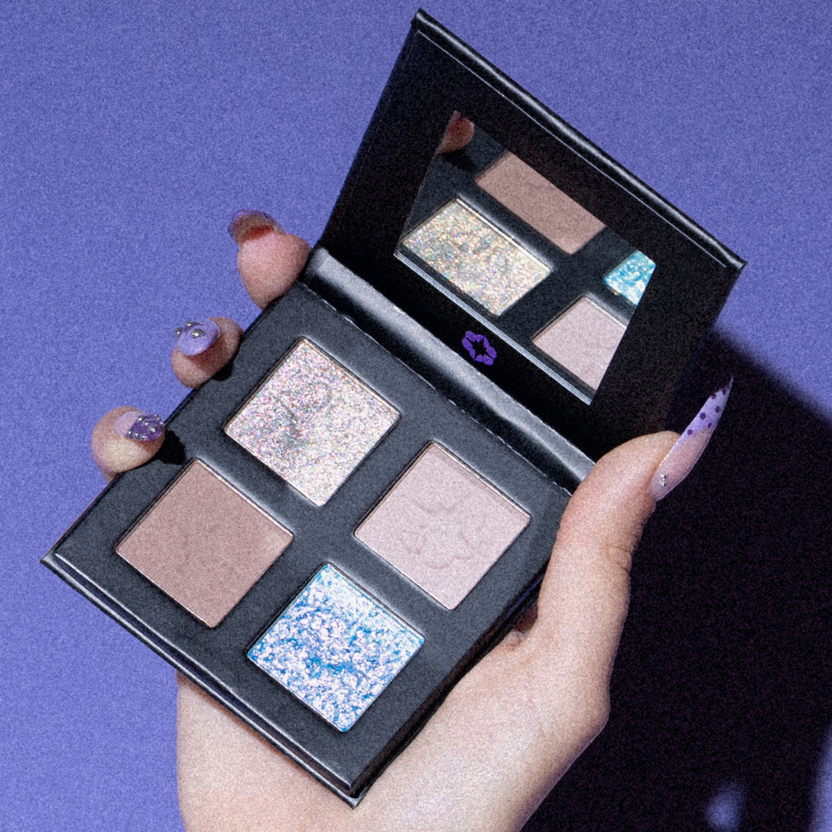 Eine Hand mit lackierten Nägeln hält die „PURISH x Lethal Palette Birthday Edition“ von Lethal Cosmetics, die auf vier Farbtöne aufgeschlagen ist, vor einem violetten Hintergrund.