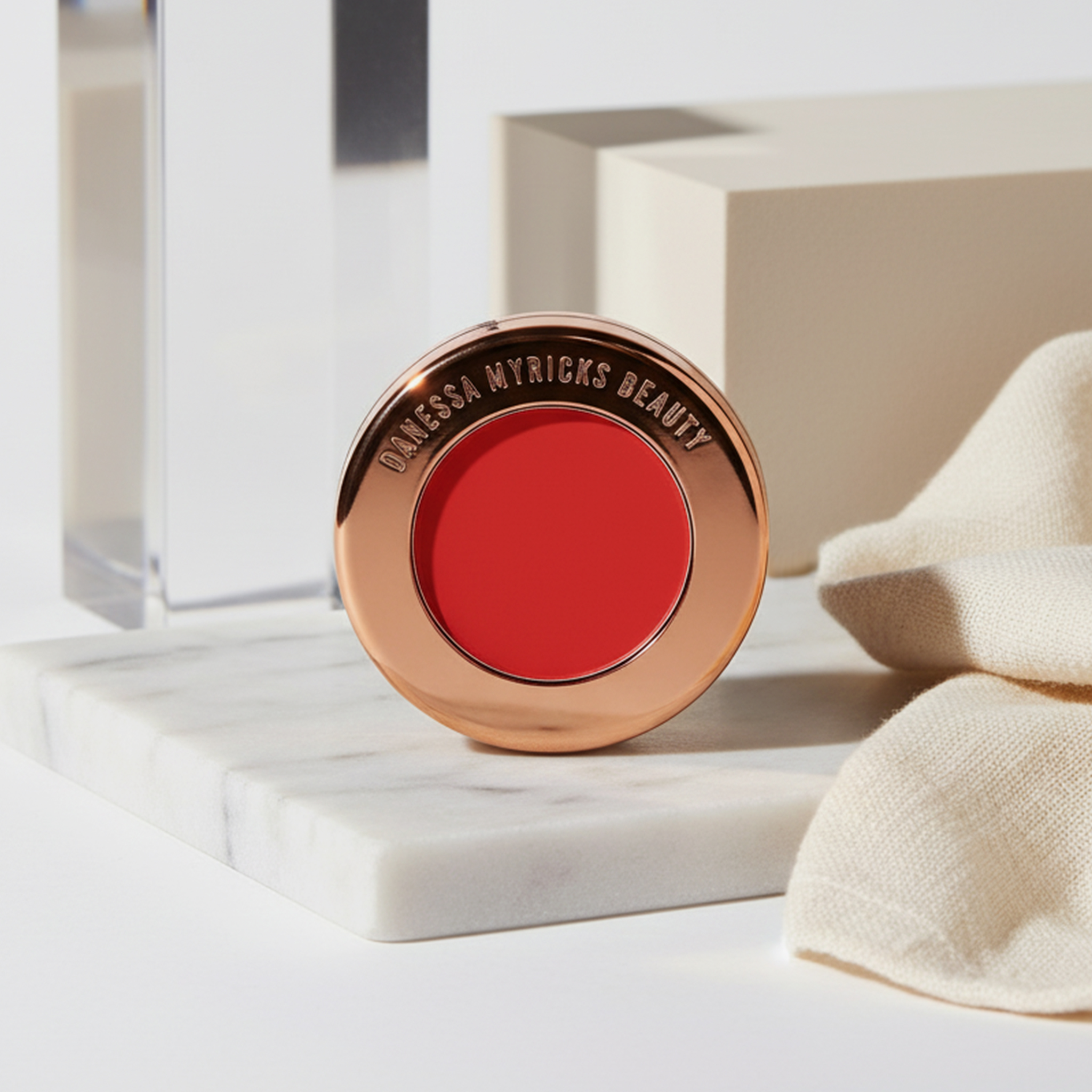 Der rote Yummy Skin Blurring Balm Powder Flushed in Red Velvet aus dem Danessa Myricks Beauty Signature Bundle, ruht auf einer Marmoroberfläche neben beigem Stoff.