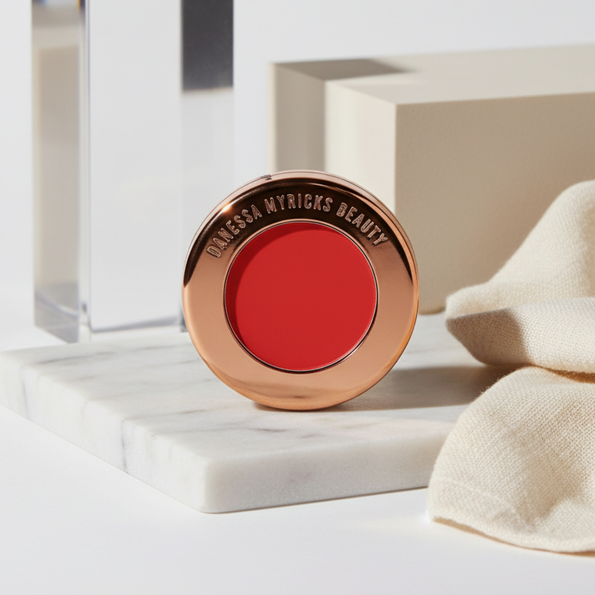 Der rote Yummy Skin Blurring Balm Powder Flushed in Red Velvet aus dem Danessa Myricks Beauty Signature Bundle, ruht auf einer Marmoroberfläche neben beigem Stoff.