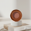 Yummy Skin Blurring Balm Powder Flushed in der Farbe Spiced Latte in rundem bronzenem Behälter auf weiß-braunem Hintergrund.