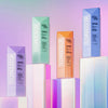 Vier Flaschen unterschiedlicher Farben des Technocolor Self Tanning Face Serum auf bunten Blöcken vor Hintergrund mit Farbverlauf.