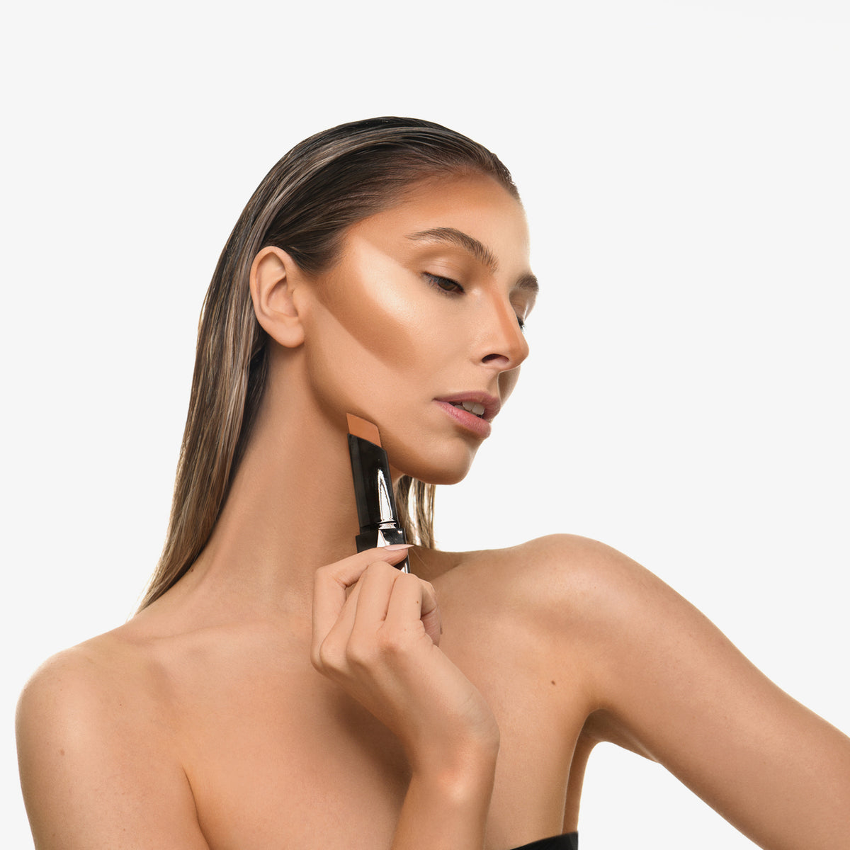 Eine Frau mit nach hinten gegelten Haaren trägt den Shapestick Bronze & Define auf ihr Gesicht auf und konturiert Wangen, Nase und Kinn.
