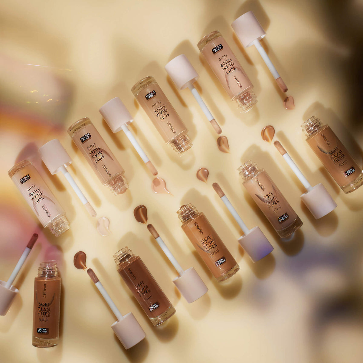 Mehrere geöffnete Flaschen und Farbmuster des Soft Glam Filter Fluid auf einer hellen gelben Oberfläche.