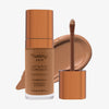 Eine transparente Flasche Yummy Skin Lift & Flex Concealer 11 neben einem bronzenen Applikator vor hellbraunem Farbmuster.