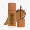Eine transparente Flasche Yummy Skin Lift & Flex Concealer 12 neben einem bronzenen Applikator vor kühl-braunem Farbmuster.