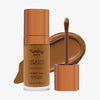 Eine transparente Flasche Yummy Skin Lift & Flex Concealer 13 neben einem bronzenen Applikator vor braunem Farbmuster.
