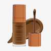 Eine transparente Flasche Yummy Skin Lift & Flex Concealer 14 neben einem bronzenen Applikator vor dunkelbraunem Farbmuster.