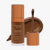 Eine transparente Flasche Yummy Skin Lift & Flex Concealer 15 neben einem bronzenen Applikator vor dunkelbraunem Farbmuster.