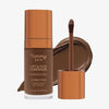 Eine transparente Flasche Yummy Skin Lift & Flex Concealer 16 neben einem bronzenen Applikator vor dunkelbraunem Farbmuster.