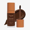 Eine transparente Flasche Yummy Skin Lift & Flex Concealer 18 neben einem bronzenen Applikator vor tiefbraunem Farbmuster.