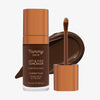 Eine transparente Flasche Yummy Skin Lift & Flex Concealer 20 neben einem bronzenen Applikator vor tiefbraunem Farbmuster.