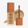 Eine transparente Flasche Yummy Skin Lift & Flex Concealer 8 neben einem bronzenen Applikator vor beigem Farbmuster.