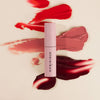 Eine Tube Pillow Soft Hydrating Lip & Cheek Stain in Pink auf cremefarbenem Hintergrund, akzentuiert mit roten, rosa und burgunderfarbenen Strichen.