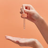 Vor einem pfirsichfarbenen Hintergrund hält eine Hand eine Pipette mit Vitamin C Glow Serum über der Anderen und ist dabei, die Flüssigkeit aufzutragen.